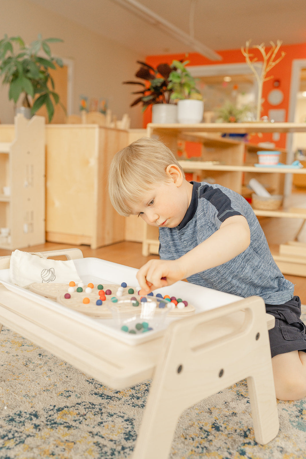 Young boy in Montessori classroom at a wooden small table working on a wooden saturn puzzle with colorful wooden balls to help hone his fine motor development. The colorful wooden balls provide nice tactile and sensory learning experience and helps with his ADHD