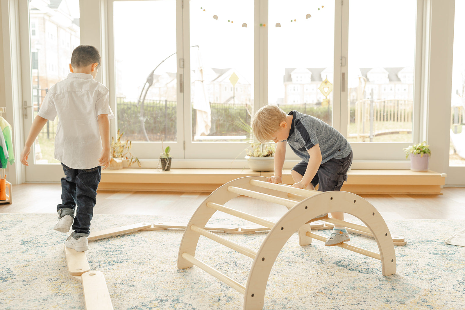 two boys playing in a Montessori classroom. One boy is climbing over the climbing arch and the other boys is balancing on the wooden balance beam next to the wooden climbing arch