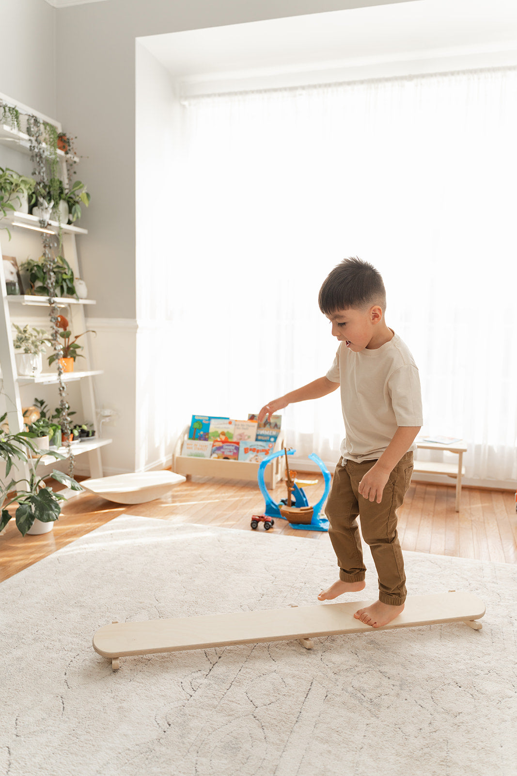 Young toddler learning balance skills on wooden wobble beam with caregiver support developing gross motor coordination and confidence. Montessori play furniture providing graduated challenge for children ages 18 months and up mastering vestibular stimulation proprioception and body awareness. Safe balance board encouraging independent movement exploration spatial orientation and physical development through open-ended rocking tilting action in home playroom classroom setting perfect for active sensory seeki