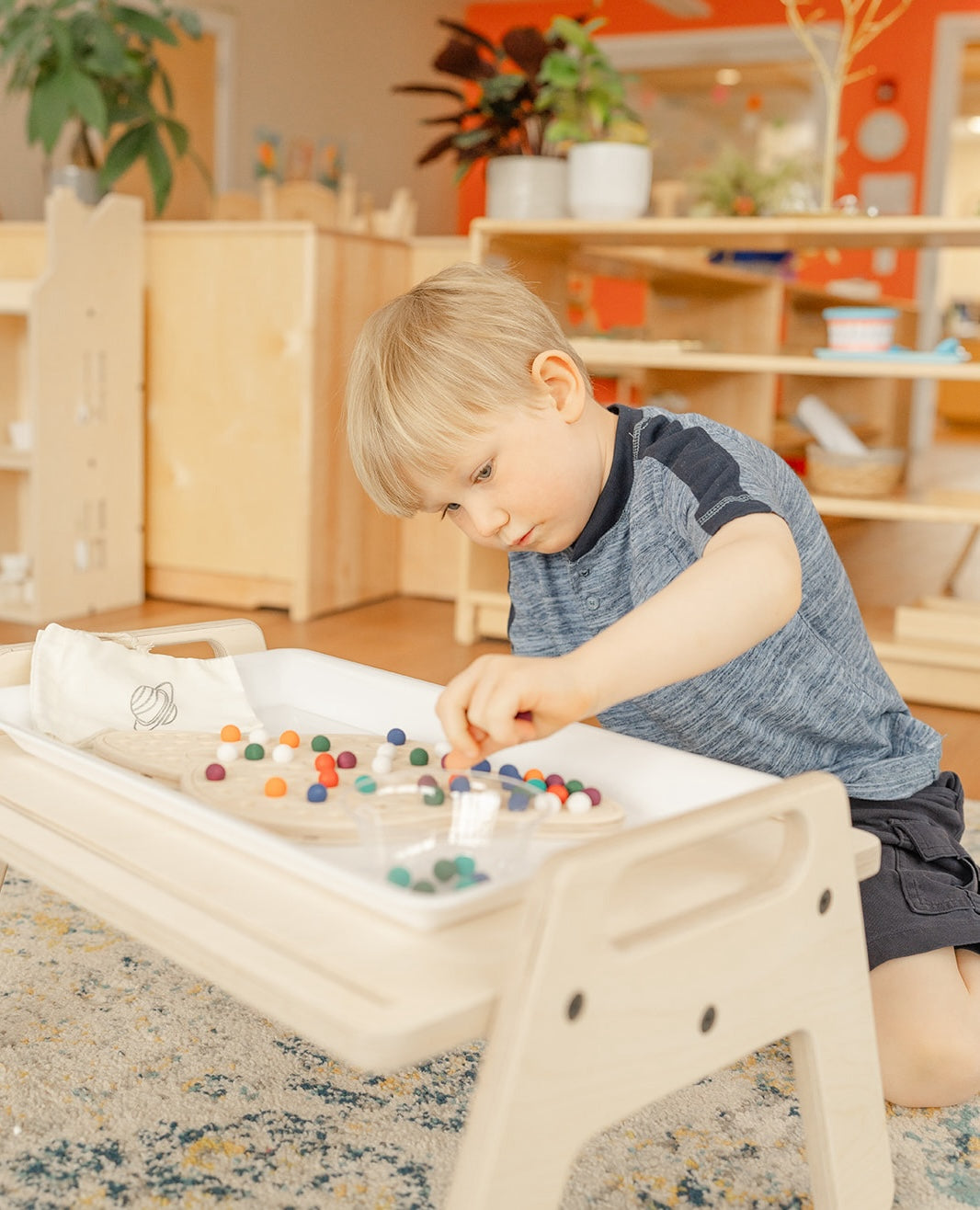 Young boy in Montessori classroom at a wooden small table working on a wooden saturn puzzle with colorful wooden balls to help hone his fine motor development. The colorful wooden balls provide nice tactile and sensory learning experience and helps with his ADHD  