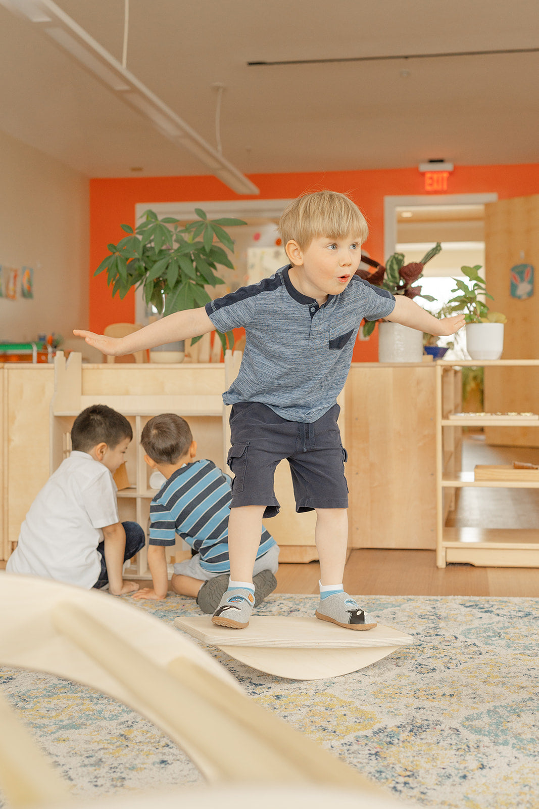A young boy in a primary Montessori Classroom on a wooden balance board challenging his gross motor and vestibular balance. 