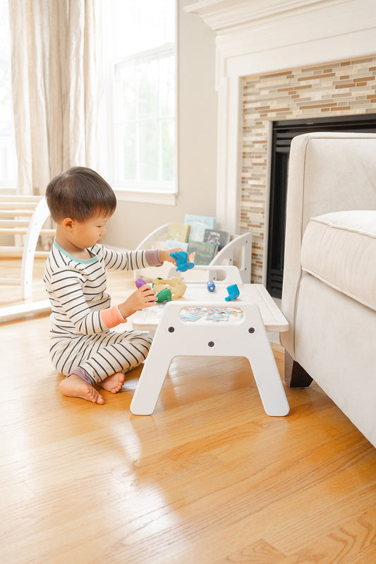 A toddler aged kid is kneeling and playing with dinosaurs on a white wooden framed small table. The table is placed in a family room and is the perfect height for babies as young as 6 months old who are learning to sit up to play and eat.