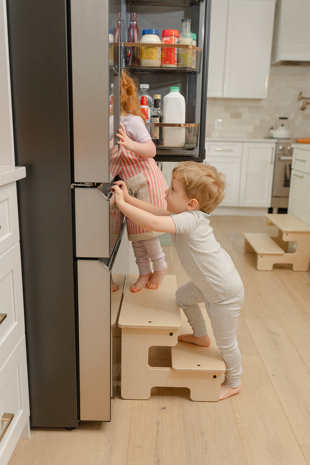 Blueberry and Third wide wooden step stool with two toddlers in mini chef aprons helping with dinner, best kitchen helper stool for little chefs ages 2 and up