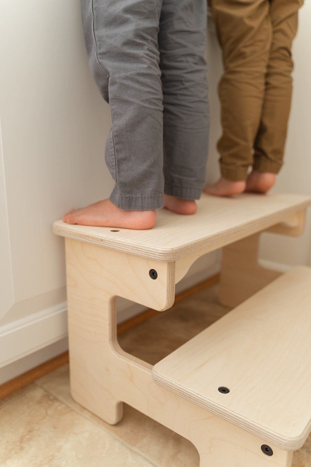 Close-up of child's feet on wooden toddler step stool showing anti-slip Baltic birch surface and safe footing during kitchen counter use, Montessori kids step stool with two solid 6-inch height steps providing stable platform for children ages 3-7 years, sturdy children's helper stool detail showing quality construction and eco-friendly water-based finish handcrafted in USA, double-wide bathroom kitchen step stool supporting 200 pounds with rounded edges for toddler safety.