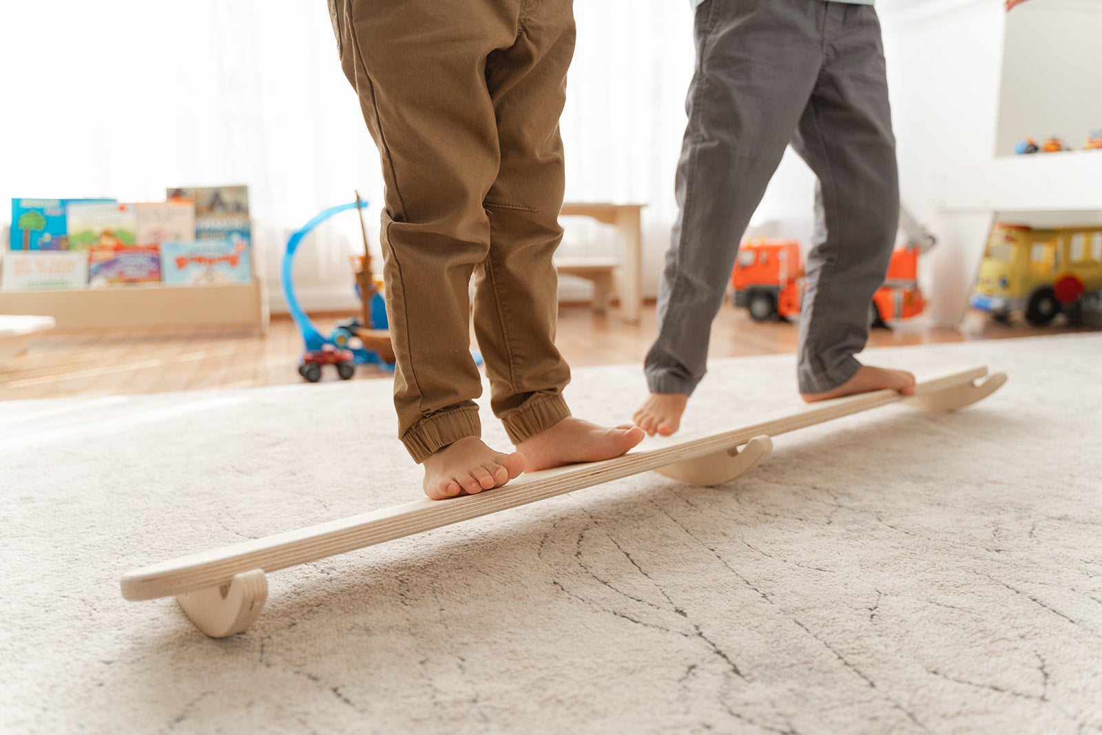 Multiple toddlers rocking together on wooden wobble beam developing social play turn-taking and shared movement experiences. Montessori balance board encouraging cooperative gross motor play vestibular stimulation and coordination development for preschool children. Open-ended play furniture supporting multiple body positions sitting standing rocking for sensory seeking kids ages 18 months to 6 years building confidence balance and physical awareness through dynamic tilting motion in playroom classroom sett