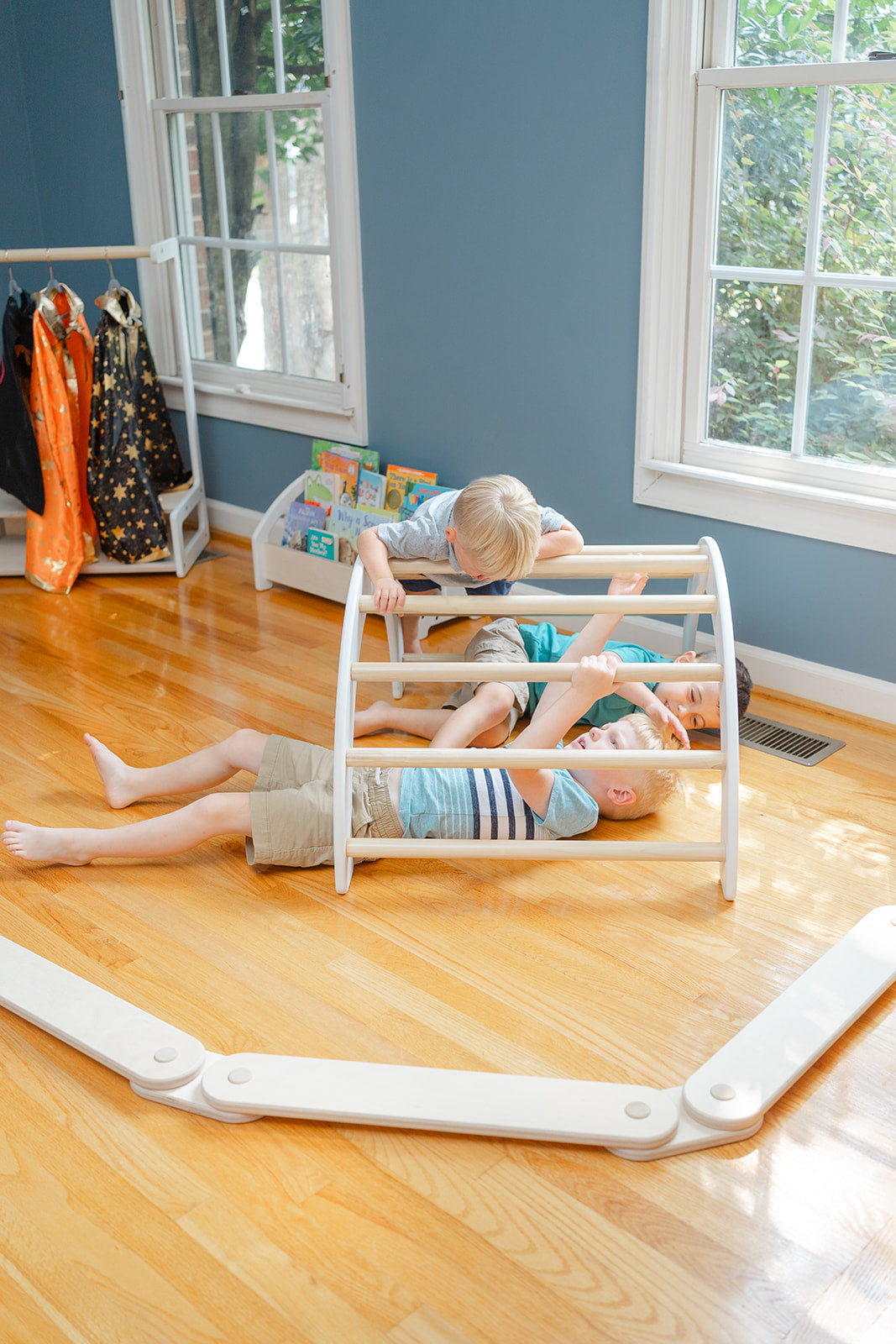 Three boys playing under wooden climbing arch using it as tunnel fort or hiding space demonstrating open ended play possibilities, Montessori Pikler arch for toddlers encouraging creative play scenarios beyond climbing including spatial awareness pretend play and social cooperation, indoor climbing toy with open-ended design allowing children to explore architecture concepts spatial relationships and collaborative storytelling while developing cognitive and social-emotional skills in playroom env
