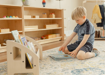 Young boy reading book on the floor next to a two-tiered floor bookshelf holding several forward facing books. The wooden bookshelf has a natural finish