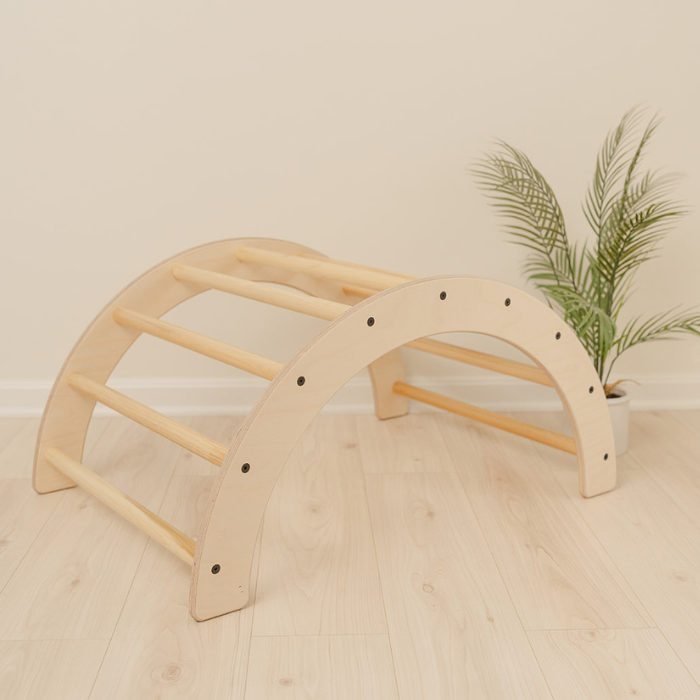 Wooden climbing arch with curved Pikler design and nine grab handles serving as versatile 2-in-1 Montessori climber and rocker for toddlers 1-3 years, indoor climbing toy for gross motor skill development through climbing crawling and balancing activities, handmade arch rocker in USA from sustainable birch plywood supporting 100 pounds perfect for playrooms bedrooms or preschool Montessori environments encouraging active play and physical exploration.