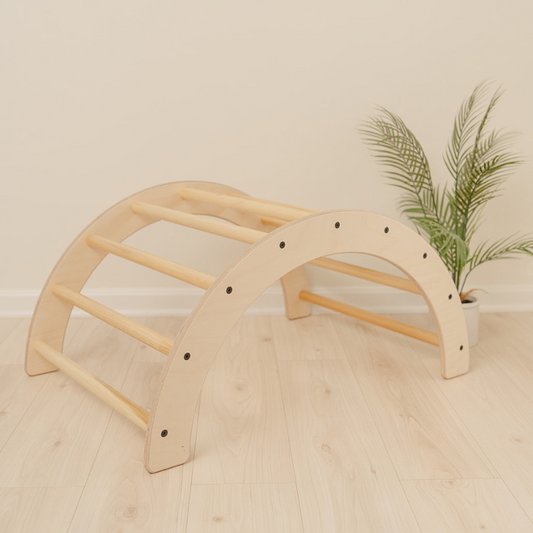 Wooden climbing arch with curved Pikler design and nine grab handles serving as versatile 2-in-1 Montessori climber and rocker for toddlers 1-3 years, indoor climbing toy for gross motor skill development through climbing crawling and balancing activities, handmade arch rocker in USA from sustainable birch plywood supporting 100 pounds perfect for playrooms bedrooms or preschool Montessori environments encouraging active play and physical exploration.