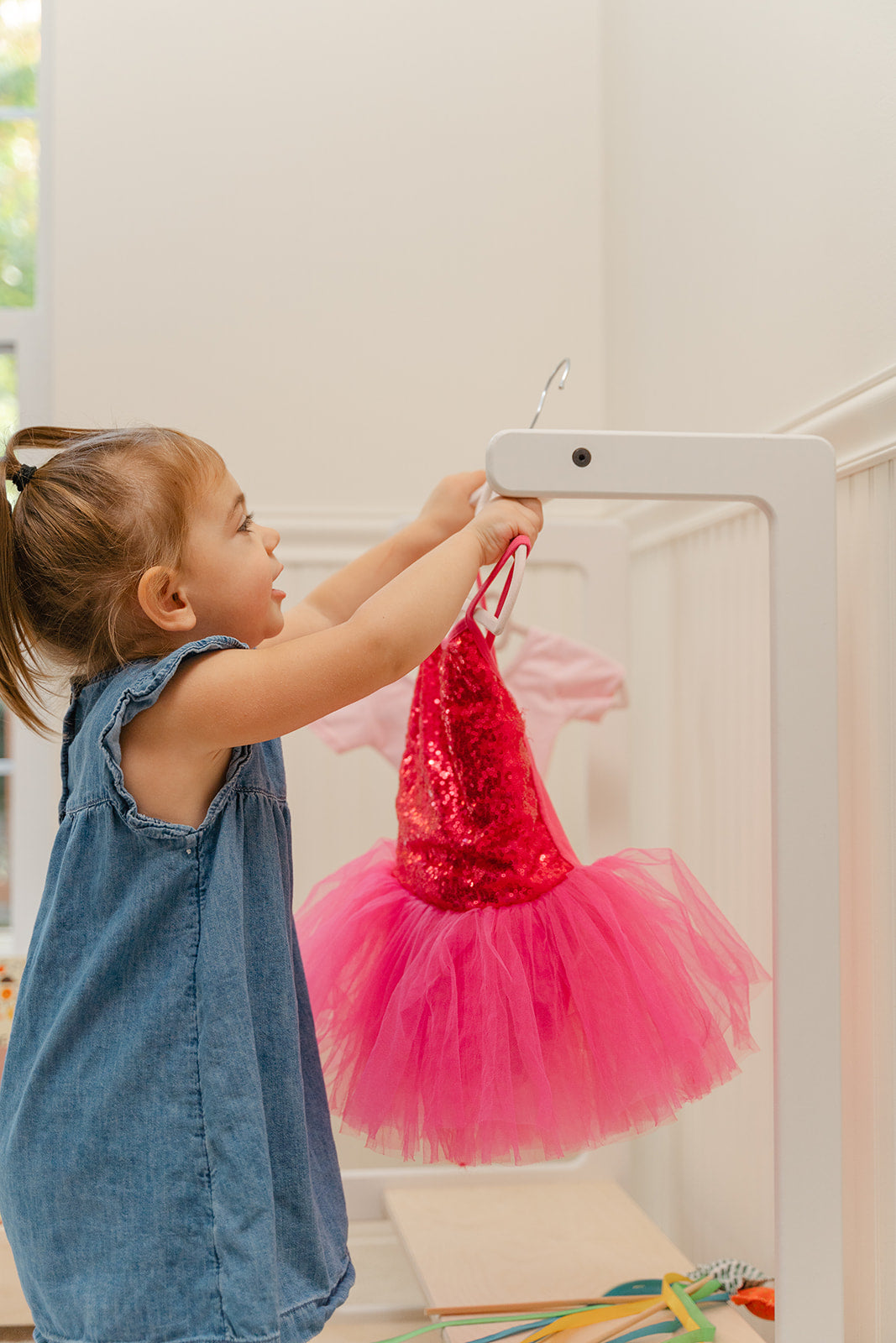 Toddler independently reaching for clothes on child's wardrobe rack at perfect child-height for autonomous dressing, Montessori kids clothing organizer empowering preschooler to select daily outfits without adult assistance, wooden children's garment rack with accessible hanging rod teaching organizational skills and morning routine independence, child-friendly clothing storage encouraging confident wardrobe choices and self-care habits.