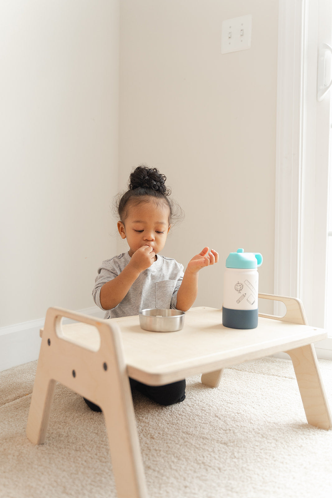Young child eating healthy snack independently at wooden toddler table on floor, portable Montessori play tray promoting self-feeding skills and mealtime independence, small activity table at perfect height for toddlers to access food and drinks without adult assistance fostering confidence and autonomy during snack time
