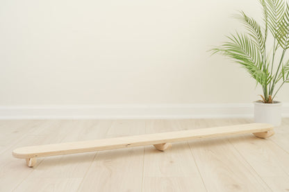 Wooden wobble beam balance board with curved base for tilting side-to-side motion developing gross motor skills vestibular system and coordination in toddlers. Montessori open-ended play furniture encouraging balance proprioception body awareness through dynamic rocking movement. USA-made birch plywood balance toy for sensory seeking children ages 18 months and up perfect for playroom classroom or home providing safe outlet for active movement and physical development experiences.