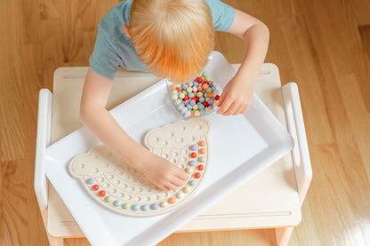 Four year old boy in a playroom playing on a small wooden table with a wooden rainbow board and colorful wooden balls. Focused on placing the colorful wooden balls in a fun design on the rainbow board.