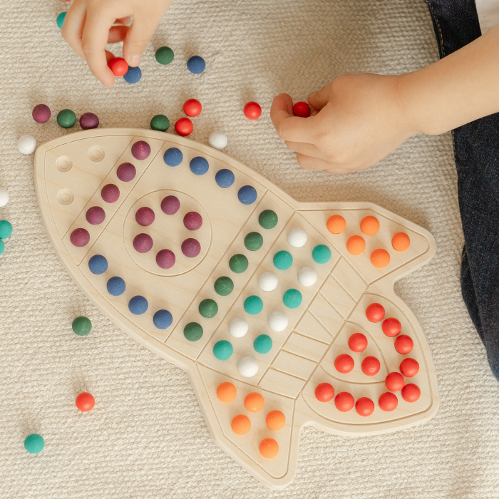 Close-up of vibrant rainbow-colored wooden pegs and balls from educational rocketship activity board showing tactile sensory elements. Colorful peg board toy with smooth wooden surfaces perfect for little hands to grasp and manipulate developing fine motor skills. Open-ended educational STEM toy featuring multicolor wooden balls encouraging color recognition sorting and pattern creation for preschoolers ages 3-6 years learning through hands-on play.
