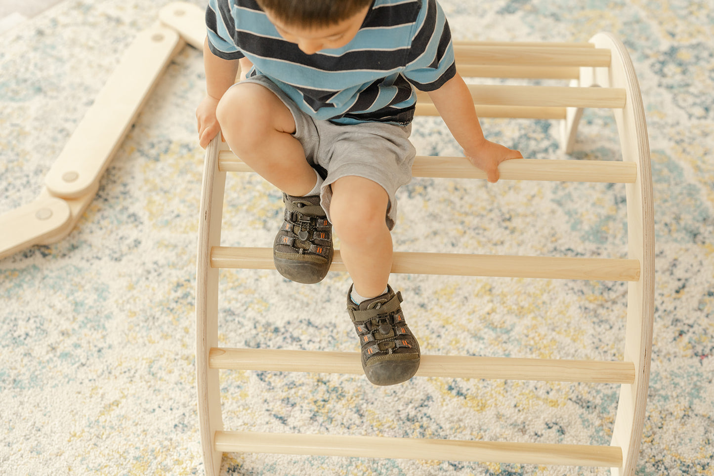 Preschool boy climbing over natural-finished wooden Pikler arch using gross motor skills demonstrating age-appropriate physical challenge for older toddlers and preschoolers, Montessori climbing arch with clear finish showcasing beautiful birch wood grain while providing climbing structure for children developing advanced coordination balance and risk assessment abilities, handcrafted indoor climbing toy from USA supporting physical development confidence building and active play for children ages 3-6 years