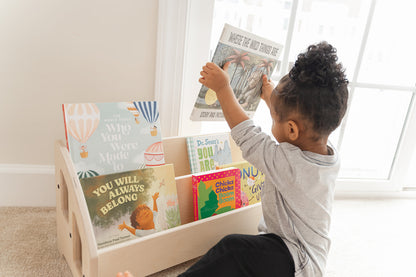 Young toddler independently selecting picture book from Montessori front-facing bookshelf demonstrating how forward-facing book display with covers visible encourages early literacy and reading independence in children ages 1-6 years old, low-height wooden kids bookshelf allows pre-readers to easily identify favorite books by recognizing colorful covers rather than text spines promoting autonomous book choices, child-sized bookshelf provides heirloom-quality nursery furniture handcrafted in USA 