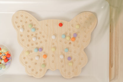 A wooden activity board designed like a butterfly, with colorful beads scattered around it, placed on a white surface with a palm leaf in the background.