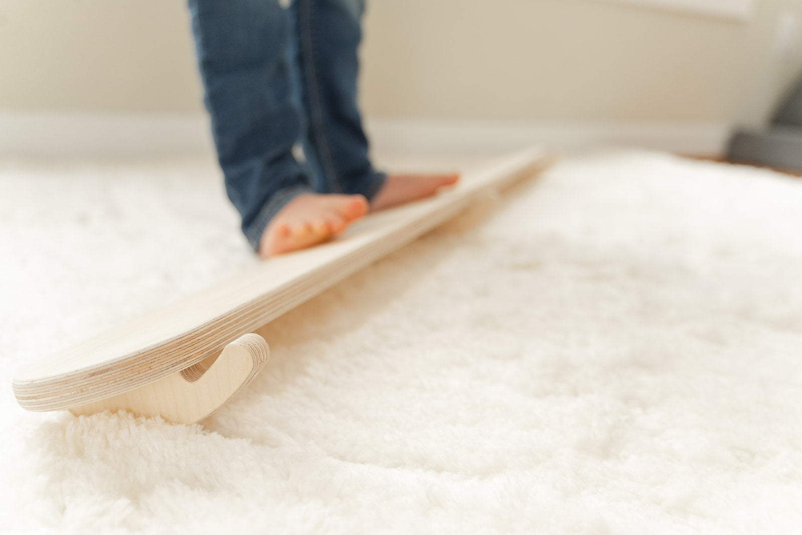 Close-up of wooden wobble beam tilting side-to-side showing unique balance challenge for toddlers developing vestibular system. Curved base design allows dynamic rocking motion stimulating proprioception body control and spatial orientation for sensory seeking children. Montessori balance board with smooth birch plywood construction providing safe challenging activity for gross motor development coordination and core strengthening through open-ended active play experiences for ages 18 months plus.