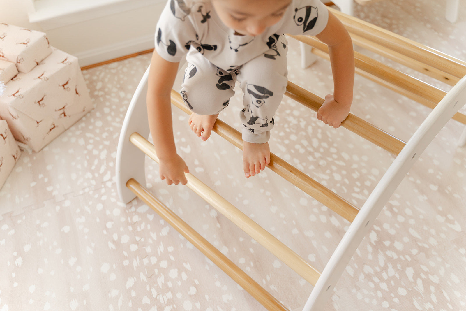 Aerial view of child climbing over Montessori arch climber showing nine rounded wooden handles perfect for toddler hand grips, white-finished climbing arch for toddlers with natural birch handles promoting hand-eye coordination and upper body strength, versatile indoor climbing toy encouraging children to explore different climbing routes and movements developing gross motor skills balance confidence and physical literacy through open-ended play.