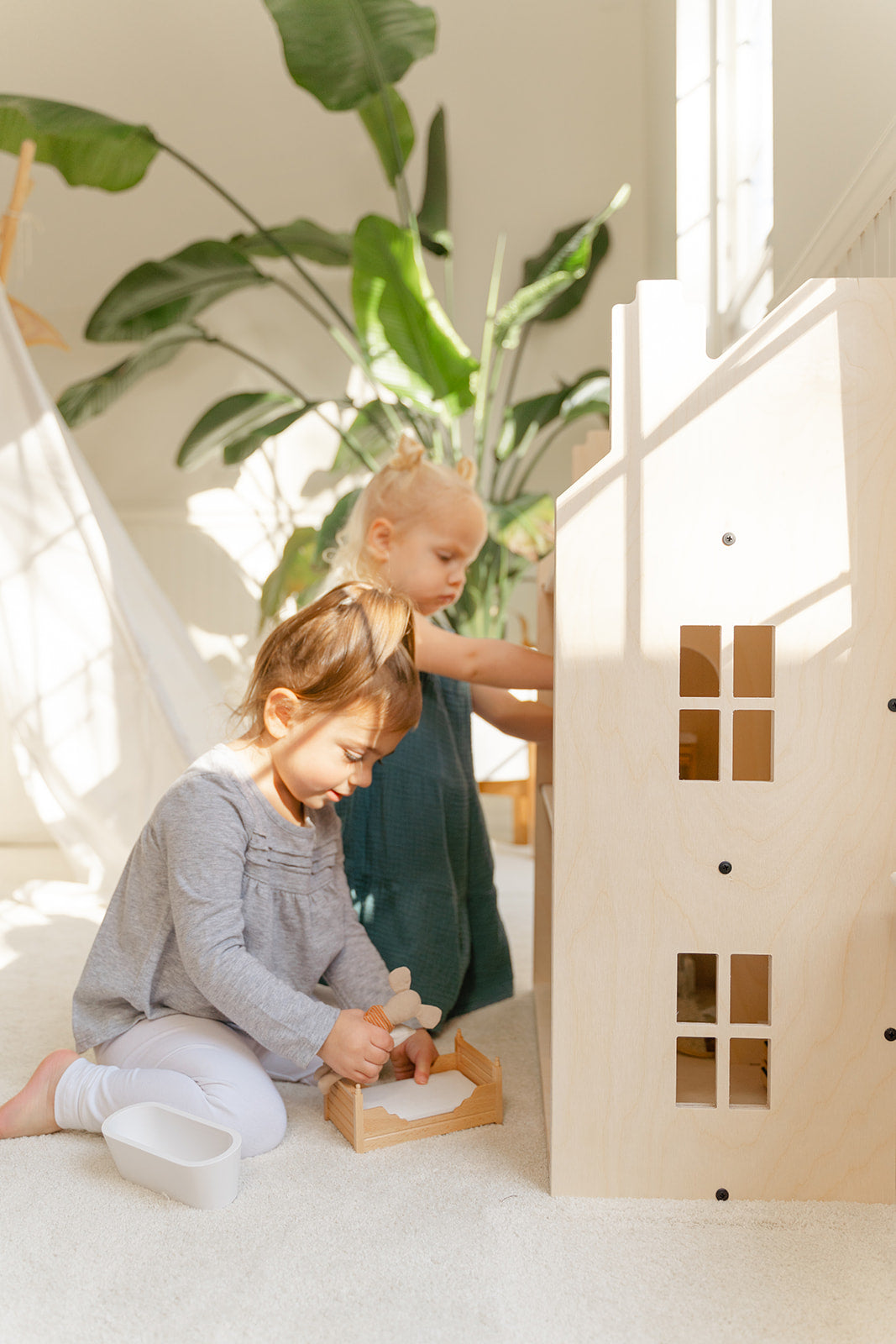 Young child engaging in imaginative pretend play with large wooden dollhouse arranging miniature furniture and dolls in rooms, life-size 3-story doll house at perfect height for kids to access all levels encouraging fine motor skills and creativity, wooden playhouse fostering storytelling abilities social-emotional development and problem-solving through open-ended play for preschool and elementary-aged children