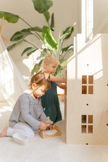 Young child engaging in imaginative pretend play with large wooden dollhouse arranging miniature furniture and dolls in rooms, life-size 3-story doll house at perfect height for kids to access all levels encouraging fine motor skills and creativity, wooden playhouse fostering storytelling abilities social-emotional development and problem-solving through open-ended play for preschool and elementary-aged children