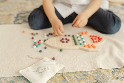 Young child using wooden tongs to pick up colorful balls and place them on rocketship activity board developing pincer grasp and hand strength. Educational fine motor skills toy with included tongs promoting proper grip development and hand-eye coordination essential for writing readiness. Montessori sensory peg board activity encouraging concentration patience and self-directed learning for preschool children ages 3-6 years through engaging tactile manipulation and STEM play.