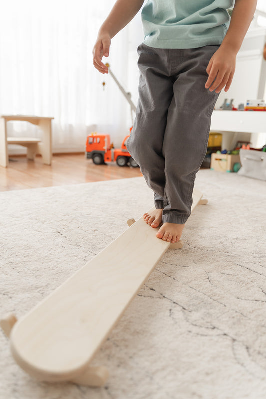 Toddler balancing on wooden wobble beam learning coordination and body awareness through vestibular stimulation. Montessori balance board developing gross motor skills concentration and spatial awareness for young children ages 18 months to 6 years. Open-ended play furniture encouraging proprioception development core strength and confidence through dynamic tilting movement. Wooden sensory toy providing safe outlet for active toddlers to rock sway and balance in playroom environment.